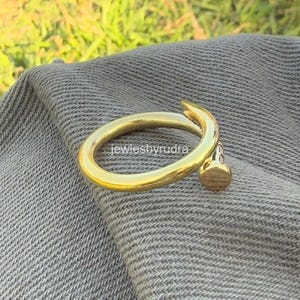 May include: A gold-coloured ring in the shape of a nail, with a rounded head and a curved shank. The ring is displayed on a grey fabric surface. The text "jewlesbyrudra" is visible on the ring.