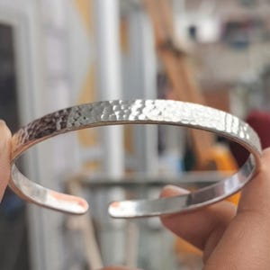 May include: A silver-colored, open cuff bracelet with a hammered texture. The bracelet is held open, revealing the inner surface. The metal has a slightly reflective quality, and the overall design is simple and elegant.