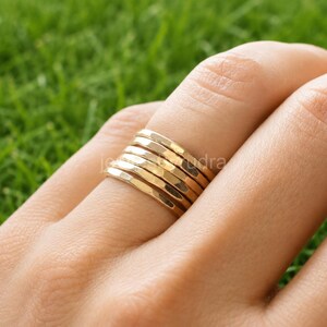 May include: A set of five stacked gold-coloured rings with a hammered, textured finish. Each ring features a flat, polished surface with subtle indentations that catch the light, creating a reflective appearance across the band stack on the finger.