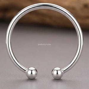 Op de afbeelding: Een gepolijste, zilverkleurige open armband met een gladde, cirkelvormige band die eindigt in twee bolvormige uiteinden. In het midden van de open ruimte staat de tekst "jewlesbyrudra" in een klein, duidelijk schreefloos lettertype.