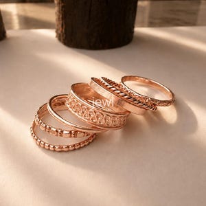 Könnte beinhalten: Ein Set aus sechs roségoldfarbenen Ringen, die übereinander gestapelt sind und verschiedene Texturen aufweisen, darunter ein Perlenband, ein glattes Band, ein gedrehtes Seilmuster und ein Band mit geprägtem Wellenmuster, mit dem Wort "jewl" in der Mitte.