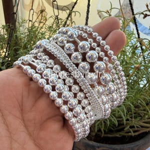 May include: A selection of silver bracelets, each with a unique design of round beads. The bracelets are held in a hand, highlighting the different textures and sizes. The beads reflect light, creating a bright, shiny appearance.