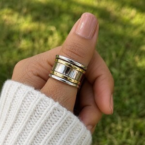 May include: A wide band ring featuring a multi-layered design with alternating polished silver-tone and gold-tone metal bands. The ring consists of a central wide silver band flanked by two thinner gold bands and two outer silver bands, creating a stacked look.