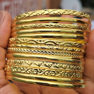 May include: A stack of gold bangles with intricate floral and geometric patterns. The bangles are held in a hand, showcasing their polished, reflective surface. The jewelry appears to be made of a shiny, yellow metal.