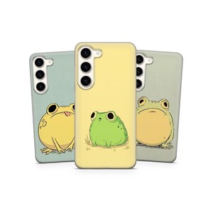 May include: Three phone cases featuring frog designs. The cases are in pale green and yellow. One case shows a yellow frog with its tongue out, another a green frog, and the third a yellow frog with a neutral expression. The cases have a camera array.