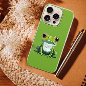 May include: A green phone case featuring a cartoon frog with large red eyes. The case is on a brown surface with a pen and a spiral notebook. The frog design is detailed with white and green accents.