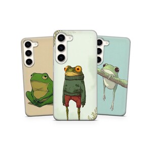 May include: Three phone cases with frog illustrations. One case features a green frog sitting, another shows a frog wearing a sweater and red pants, and the third depicts a frog on a branch. The cases have a glossy finish.