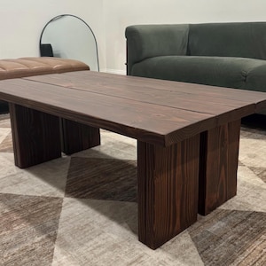 May include: A dark brown wooden coffee table with a rectangular top and four sturdy, block-style legs. The table is made of solid wood and has a natural wood grain finish. The table is a centerpiece for a living room.