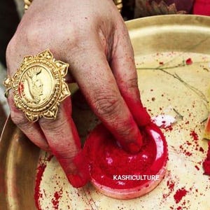 May include: A gold ring with an intricate design is worn on a finger. A red powder container and a small box are also visible. The image depicts a cultural or religious ceremony.