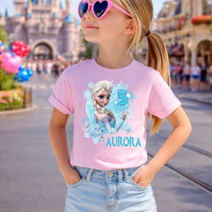 Pu&ograve; includere: Una bambina indossa una maglietta rosa con un personaggio dei cartoni animati e il nome "AURORA". La maglietta presenta anche il numero "5". Indossa pantaloncini di jeans e occhiali da sole rosa a forma di cuore.
