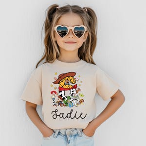 K&ouml;nnte beinhalten: Beiges T-Shirt mit farbenfroher Grafik von Toy Story-Charakteren und dem Namen "Sadie" in Schreibschrift. Das Kind tr&auml;gt herzf&ouml;rmige Sonnenbrillen.