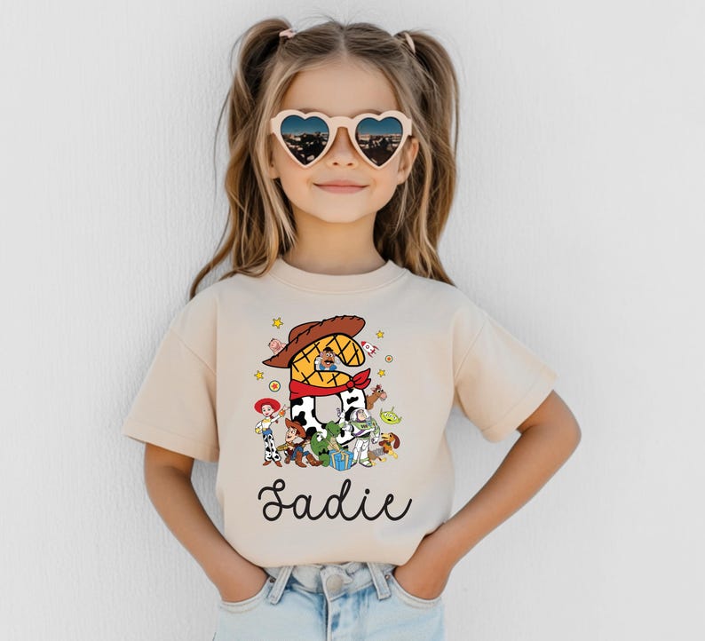 K&ouml;nnte beinhalten: Beiges T-Shirt mit farbenfroher Grafik von Toy Story-Charakteren und dem Namen "Sadie" in Schreibschrift. Das Kind tr&auml;gt herzf&ouml;rmige Sonnenbrillen.