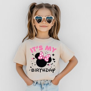 May include: A beige t-shirt featuring a black Minnie Mouse head with a pink bow and the text "IT'S MY BIRTHDAY" in pink and black. The shirt is worn by a young girl wearing heart-shaped sunglasses.