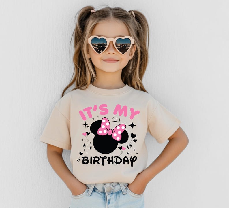 May include: A beige t-shirt featuring a black Minnie Mouse head with a pink bow and the text "IT'S MY BIRTHDAY" in pink and black. The shirt is worn by a young girl wearing heart-shaped sunglasses.