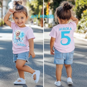 Pu&ograve; includere: Una bambina indossa una maglietta rosa chiaro con un personaggio dei cartoni animati, il nome "Aurora" e il numero "5". Indossa anche pantaloncini di jeans e scarpe bianche. La foto &egrave; stata scattata all'aperto.