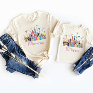 May include: Two cream-colored t-shirts with a colorful castle graphic and the words "Mommy" and "Sister" in pastel colors. The shirts are paired with blue denim jeans.