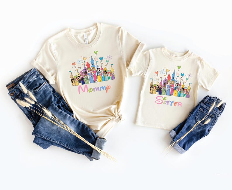 May include: Two cream-colored t-shirts with a colorful castle graphic and the words "Mommy" and "Sister" in pastel colors. The shirts are paired with blue denim jeans.