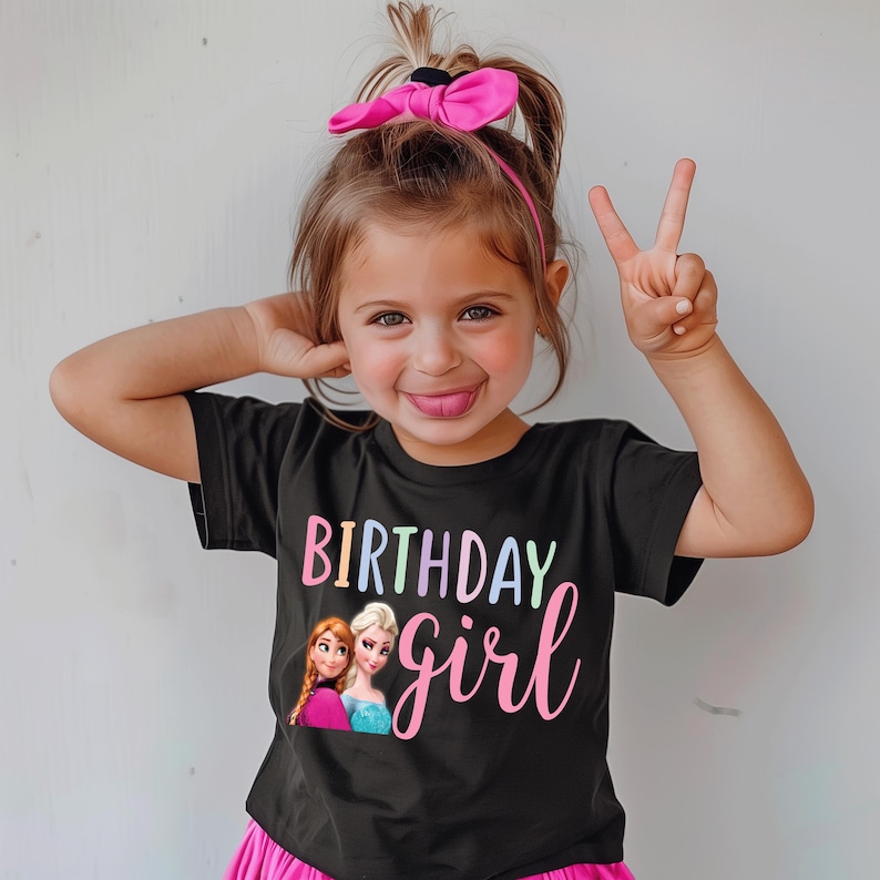 K&ouml;nnte beinhalten: Schwarzes T-Shirt mit den Worten "BIRTHDAY girl" in farbenfroher Schrift und einem Bild von zwei Zeichentrickfiguren. Das M&auml;dchen auf dem Bild tr&auml;gt eine rosa Schleife und einen Rock und macht ein Friedenszeichen mit ihren Fingern.