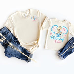 May include: Two cream-colored t-shirts and two pairs of blue denim jeans. The larger shirt features a small graphic of a blonde character and the text "Mimi". The smaller shirt displays a larger graphic of the same character with the text "BIRTHDAY GIRL".