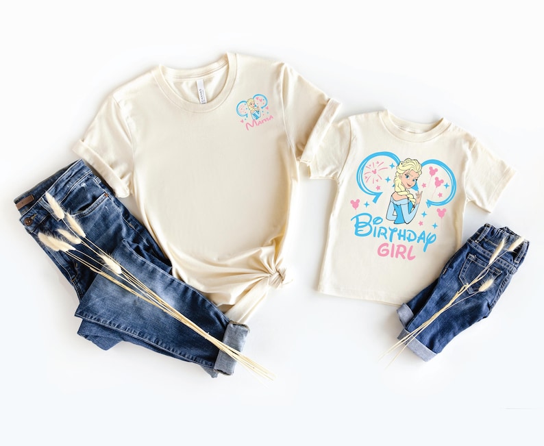 May include: Two cream-colored t-shirts and two pairs of blue denim jeans. The larger shirt features a small graphic of a blonde character and the text "Mimi". The smaller shirt displays a larger graphic of the same character with the text "BIRTHDAY GIRL".