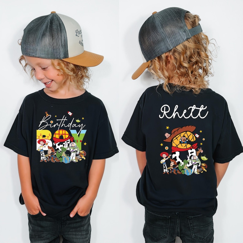 May include: Black t-shirt featuring a colorful graphic of characters and the text "Birthday BOY". The back of the shirt displays the name "Rhett" and a matching character design. The child is wearing a gray and tan baseball cap.