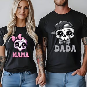 May include: Two black t-shirts featuring graphic skull illustrations. The left shirt displays a skull with a pink polka-dot bow and the text MAMA in pink. The right shirt features a skull wearing a black baseball cap and bow tie with the text DADA.