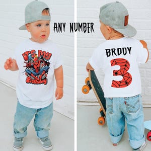 May include: A child in a white t-shirt featuring a Spider-Man design and the words "It's My Birthday". The child is also wearing blue jeans and a grey baseball cap. The back of the shirt has the name "Broddy" and the number "3".