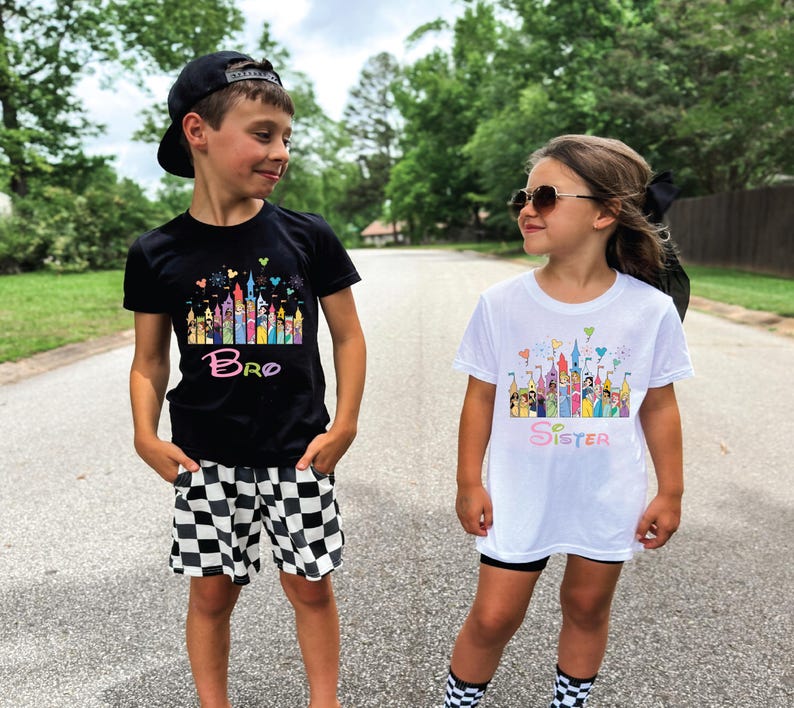 May include: Two children wearing matching graphic t-shirts. One shirt is black with the word "Bro" and a colorful castle design. The other shirt is white with the word "Sister" and a similar castle design. The boy wears checkered shorts and a black cap.