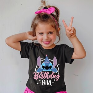 May include: A black t-shirt featuring a cartoon character with large ears and a party hat. The text "Birthday Girl" is written in pink script below the character. The girl is wearing a pink bow and pink skirt.