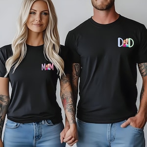 K&ouml;nnte beinhalten: Schwarze T-Shirts mit passenden Designs. Ein Shirt hat das Wort "MOM" in Pink und Lila, das andere "DAD" in Blau und Gr&uuml;n. Beide Shirts haben ein kleines Bild einer Familie. Die Personen tragen blaue Jeans.
