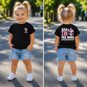 May include: A black t-shirt featuring a small pink and white skull graphic with a bow and the name ARIA on the front left chest. The back displays the text BAD TWO THE BONE in large white letters with a pink skull and a black and white checkered border.