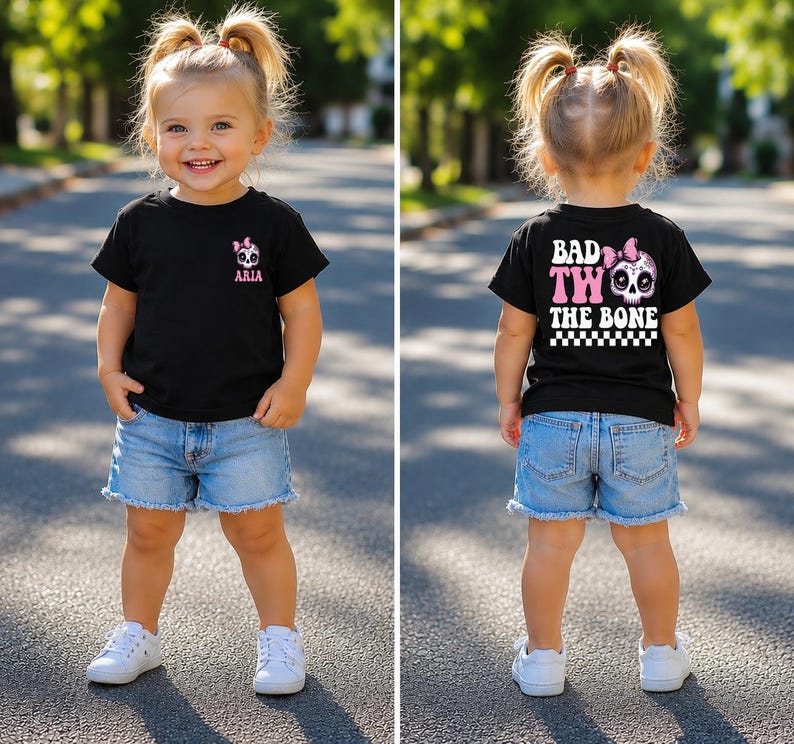 May include: A black t-shirt featuring a small pink and white skull graphic with a bow and the name ARIA on the front left chest. The back displays the text BAD TWO THE BONE in large white letters with a pink skull and a black and white checkered border.