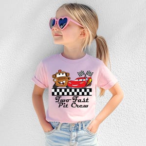 Puede incluir: Una camiseta rosa claro con un gr&aacute;fico de dos coches de dibujos animados y el texto "Two Fast Pit Crew" en negro. La camiseta la lleva una persona joven con gafas de sol en forma de coraz&oacute;n.