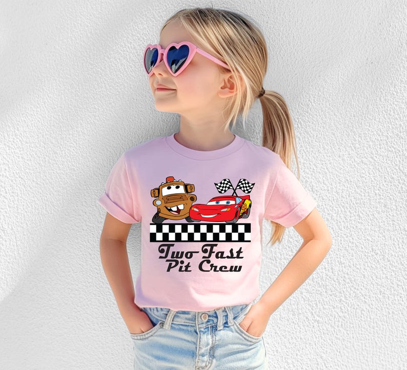 Puede incluir: Una camiseta rosa claro con un gr&aacute;fico de dos coches de dibujos animados y el texto "Two Fast Pit Crew" en negro. La camiseta la lleva una persona joven con gafas de sol en forma de coraz&oacute;n.
