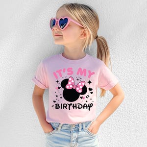 May include: A light pink t-shirt with the text "IT'S MY BIRTHDAY" and a black Minnie Mouse head with a pink polka dot bow. The girl is wearing pink heart-shaped sunglasses and blue jeans.