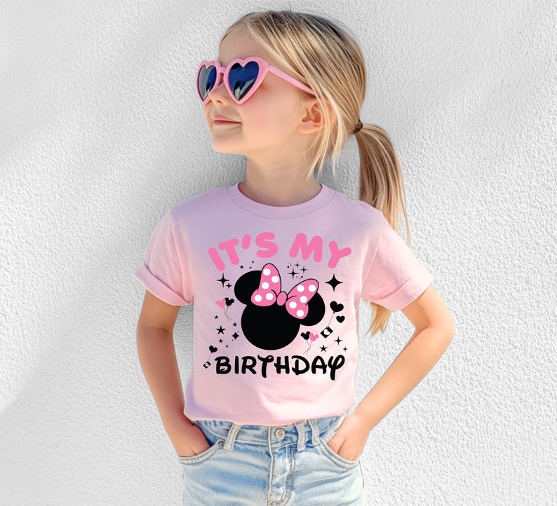 May include: A light pink t-shirt with the text "IT'S MY BIRTHDAY" and a black Minnie Mouse head with a pink polka dot bow. The girl is wearing pink heart-shaped sunglasses and blue jeans.