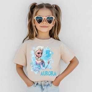 Pu&ograve; includere: Una maglietta beige con una grafica di una principessa dei cartoni animati, un numero 5 blu e il nome "AURORA" in blu. La ragazza indossa occhiali da sole a forma di cuore.