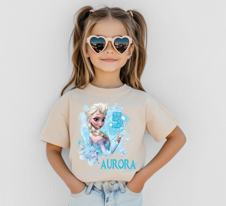 Pu&ograve; includere: Una maglietta beige con una grafica di una principessa dei cartoni animati, un numero 5 blu e il nome "AURORA" in blu. La ragazza indossa occhiali da sole a forma di cuore.