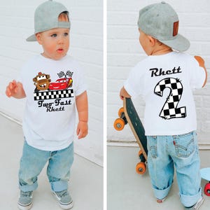 May include: White toddler t-shirt with a graphic of Mater and Lightning McQueen from the movie Cars, with the text "Two Fast Rhett". The back of the shirt has a checkered number 2 and the name "Rhett". The child is wearing blue jeans and a gray baseball cap.