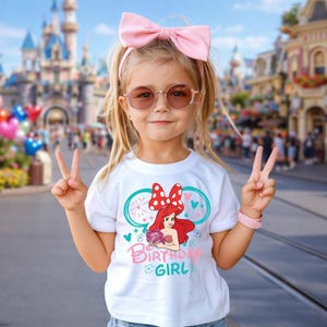 May include: A white short-sleeved t-shirt featuring a graphic of Ariel with a red bow, teal mouse ears, and the text Birthday Girl in pink and teal. The shirt is worn with a large light pink hair bow and round-framed sunglasses.