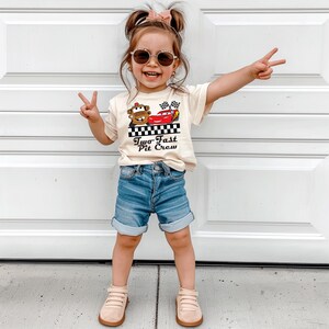 Puede incluir: Una ni&ntilde;a lleva una camiseta color crema con un dibujo de dos coches de dibujos animados y el texto "Two Fast Pit Crew". La ni&ntilde;a tambi&eacute;n lleva pantalones cortos vaqueros azules, gafas de sol y zapatos color beige.