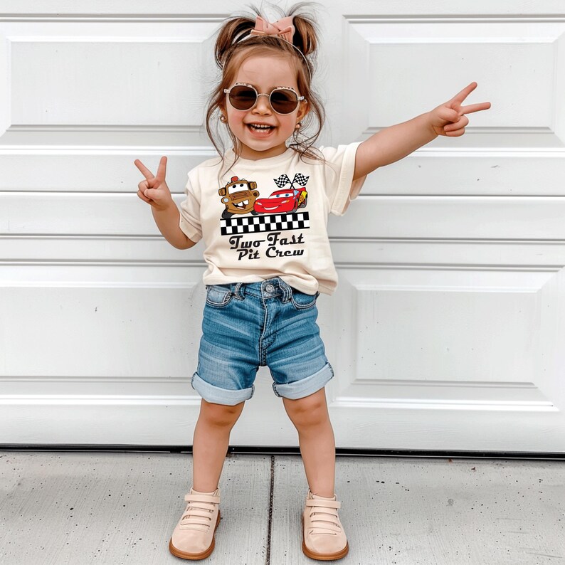 Puede incluir: Una ni&ntilde;a lleva una camiseta color crema con un dibujo de dos coches de dibujos animados y el texto "Two Fast Pit Crew". La ni&ntilde;a tambi&eacute;n lleva pantalones cortos vaqueros azules, gafas de sol y zapatos color beige.