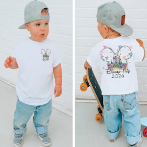 Pu&ograve; includere: Un bambino indossa una maglietta bianca e jeans azzurri. La maglietta presenta una grafica colorata a tema Disney sul davanti e sul retro, con la scritta "Disney Trip 2026". Il bambino indossa anche un berretto da baseball grigio.