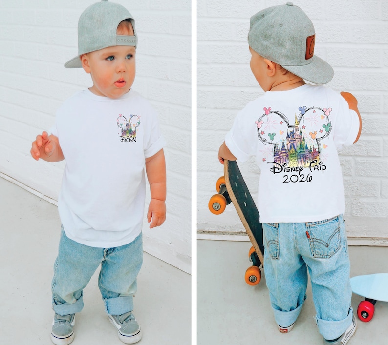 Pu&ograve; includere: Un bambino indossa una maglietta bianca e jeans azzurri. La maglietta presenta una grafica colorata a tema Disney sul davanti e sul retro, con la scritta "Disney Trip 2026". Il bambino indossa anche un berretto da baseball grigio.