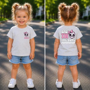May include: A light gray short-sleeve t-shirt featuring a small pink skull graphic with a bow on the front left chest with the name Aria. The back displays a large pink skull with a bow and the text Bad Two The Bone above a checkered pattern.