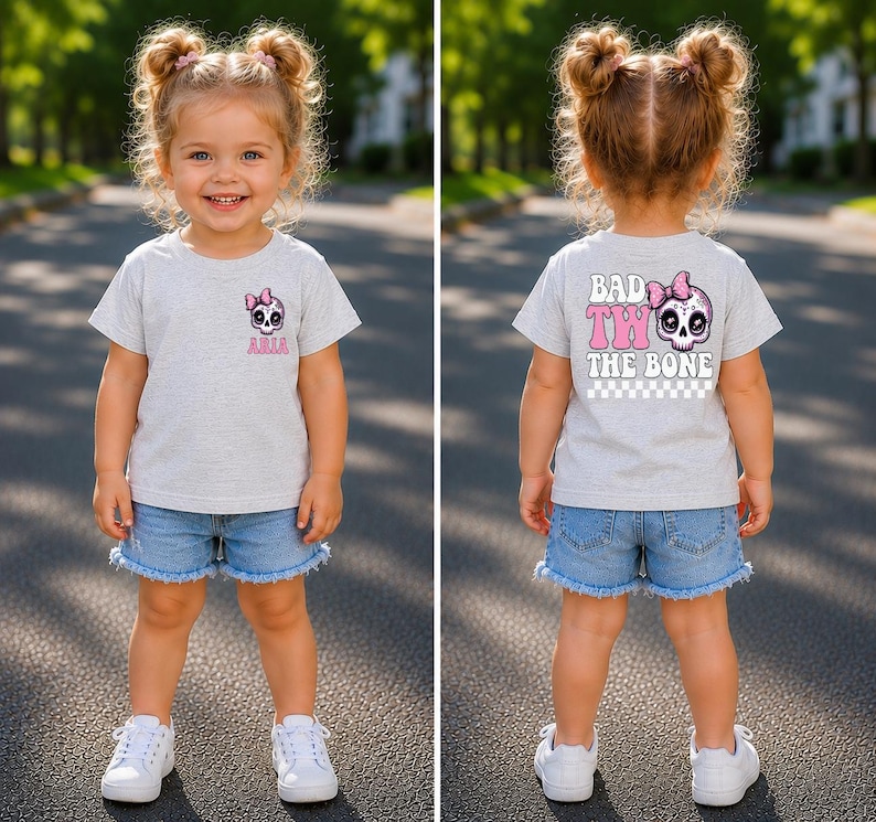May include: A light gray short-sleeve t-shirt featuring a small pink skull graphic with a bow on the front left chest with the name Aria. The back displays a large pink skull with a bow and the text Bad Two The Bone above a checkered pattern.
