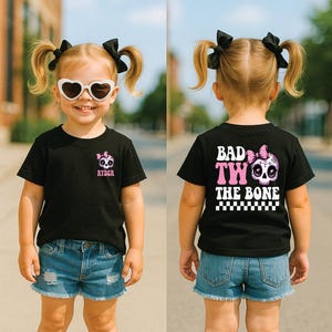 May include: A black t-shirt featuring a small pink and white skull graphic with a bow and the name Ryder on the front left chest. The back displays a large skull graphic with a pink bow, the text Bad Two The Bone, and a black and white checkerboard.
