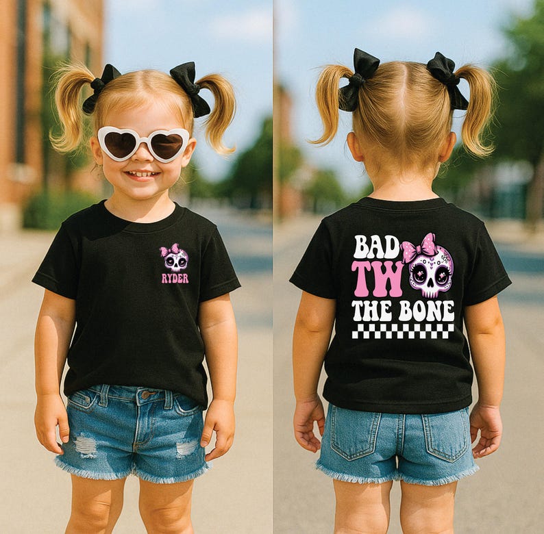 May include: A black t-shirt featuring a small pink and white skull graphic with a bow and the name Ryder on the front left chest. The back displays a large skull graphic with a pink bow, the text Bad Two The Bone, and a black and white checkerboard.