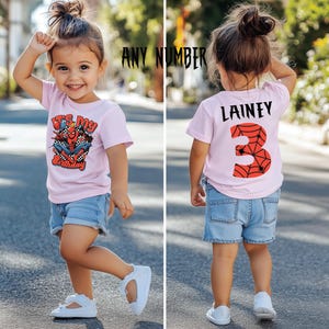 May include: A child in a light pink t-shirt and denim shorts. The front of the shirt displays a Spider-Man graphic and the text "It's My Birthday". The back of the shirt has the name "Lainey" and the number "3" with a spiderweb design.