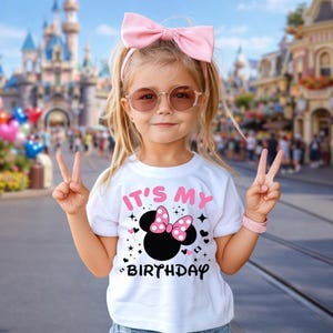 May include: A young girl wearing a white t-shirt with the text "IT'S MY BIRTHDAY" and a Minnie Mouse design. She is wearing a pink bow, sunglasses, and a pink bracelet. She is giving the peace sign.